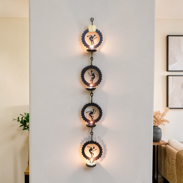 Handmade Bastar iron craft circular tea light wall hanging displayed vertically on a wall, featuring four black wrought iron circle panels with folk dancer motifs and glowing diyas