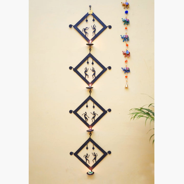 Handmade Bastar iron craft square tiles tea light wall hanging displayed vertically on a wall, featuring four black wrought iron panels with folk dancer motifs and glowing diyas