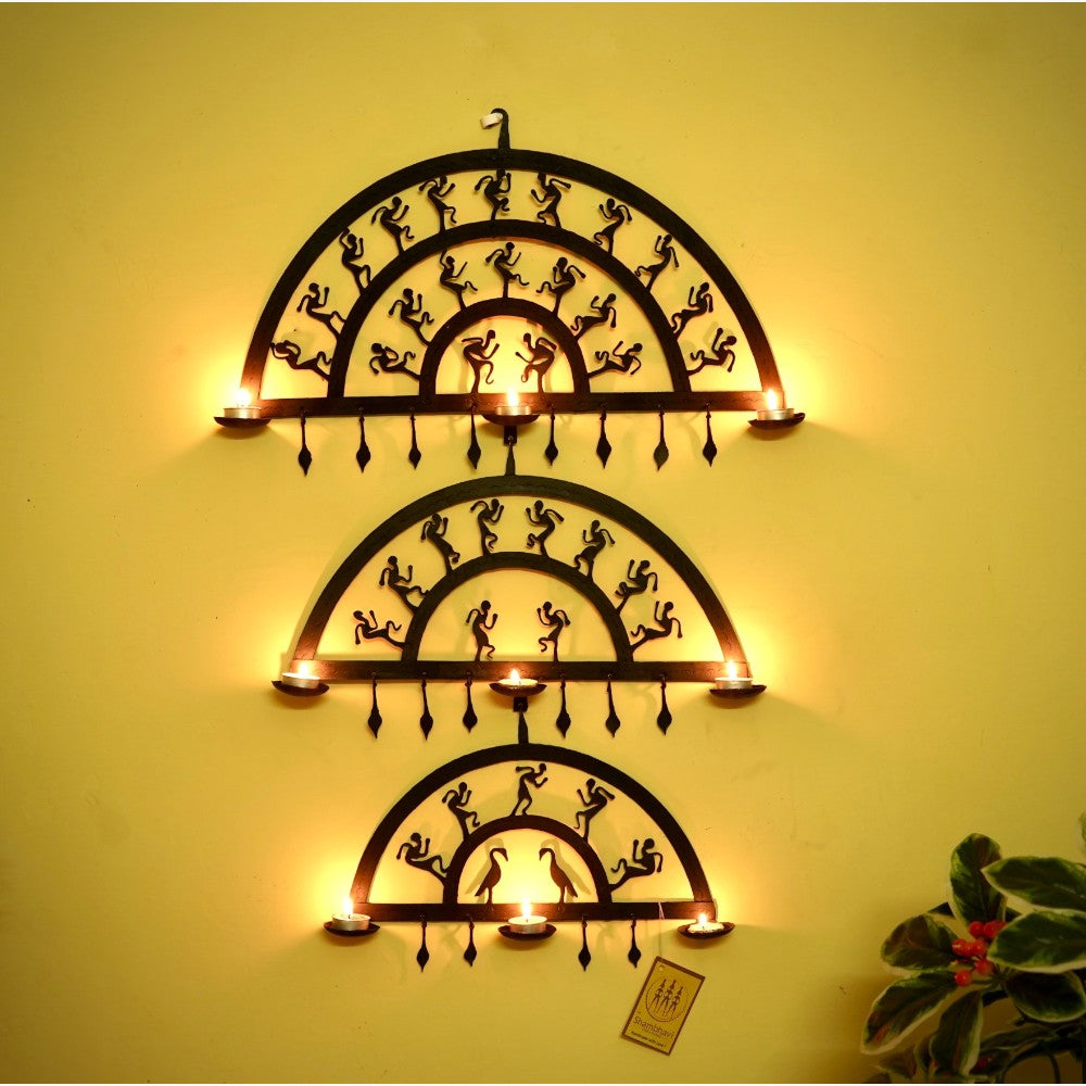 Handmade Bastar iron art half circle wall hanging displayed on a wall with lit diyas, featuring layered semicircular wrought iron frames and folk dancer motifs creating a warm ambient glow