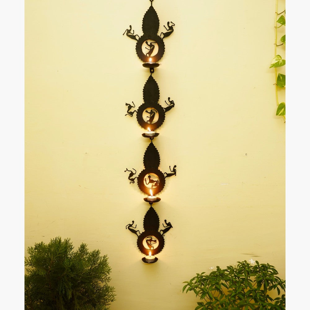 Handmade Bastar iron craft leaf design tea light wall hanging displayed vertically on a wall, featuring black wrought iron panels with folk dancer motifs and softly glowing diyas