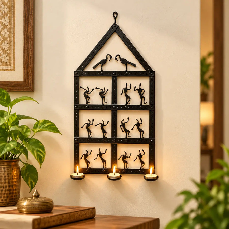 Bastar Loha Shilp wrought iron wall hanging shaped like a home, with dancing figures, two birds and three lit tealights on a cream wall