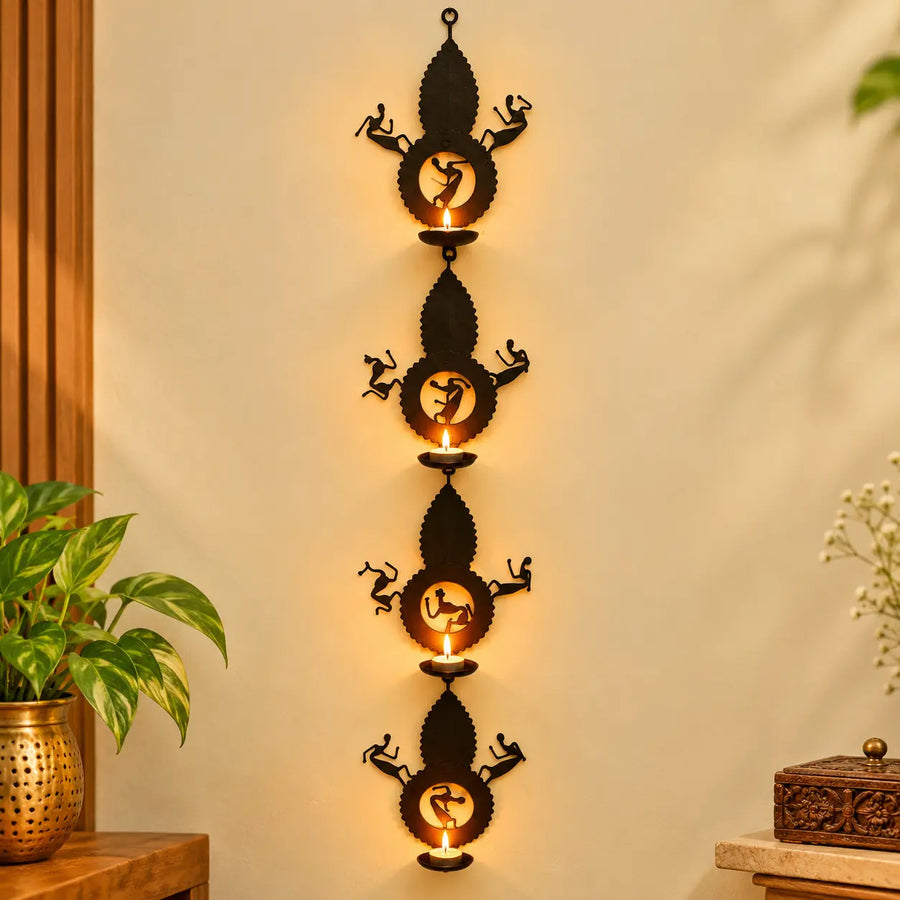 Bastar Loha Shilp wrought iron leaf wall hanging with four tea light holders and dancing figures, glowing on a cream wall