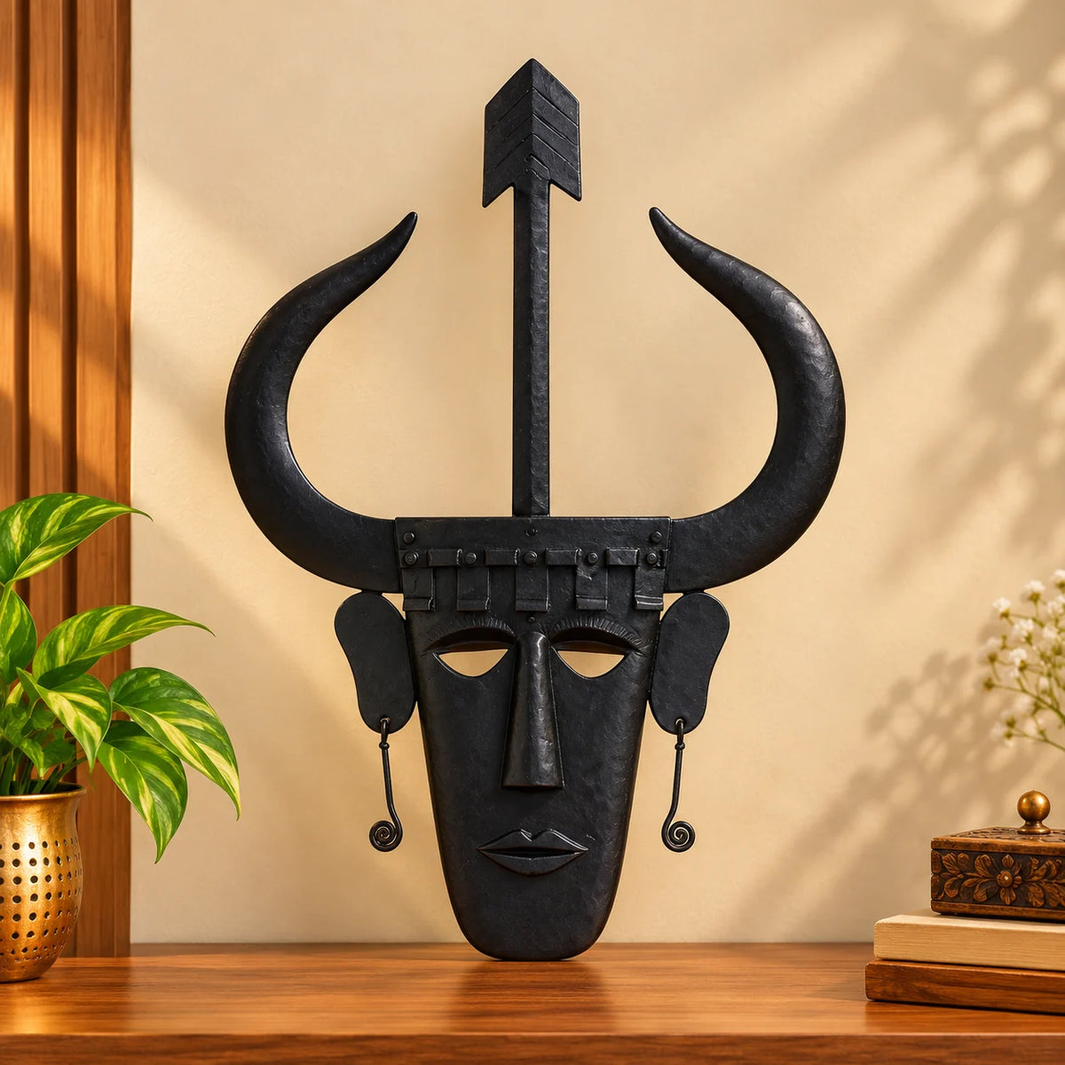 Madia wrought iron wall mask with curved horns and arrow crest, handcrafted in Bastar Loha Shilp style, on a wooden shelf